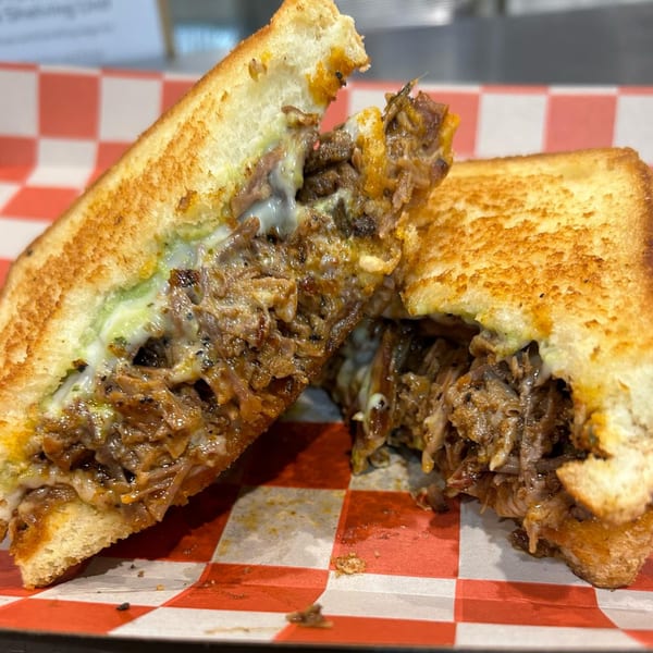 A grilled sandwich, cut in half, revealing juicy pulled pork and melted cheese on red and white checkered paper.